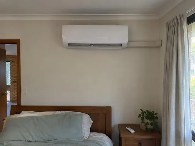 Clean split system mounted high on a bedroom wall above a bed, with curtains and a bedside table in Robina